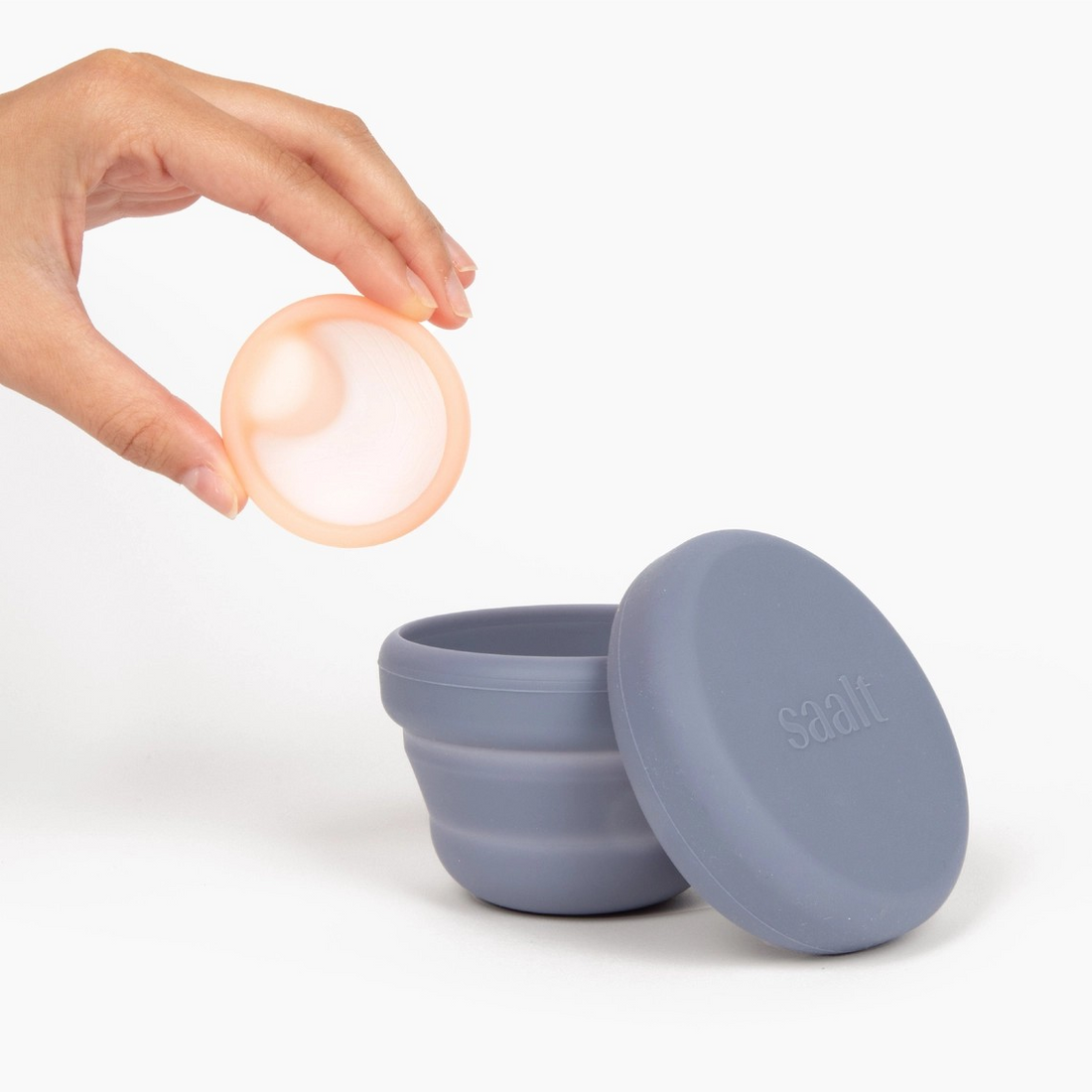 Saalt compact sanitizer in slate blue with lid removed, next to a hand holding a pink blush menstrual disc.