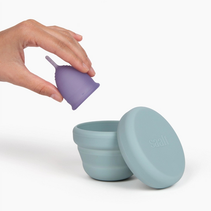 Saalt compact sanitizer in slate blue with lid removed, next to a hand holding a purple menstrual cup.